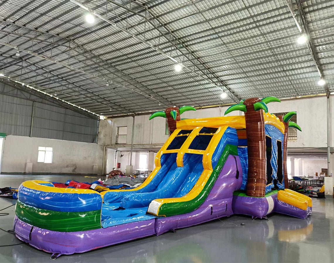 Wide-angle view of Goombay Splash Dual Lane Commercial Inflatable Combo in a spacious warehouse. The inflatable features dual curved blue slides, palm tree pillars, a yellow and blue bounce house structure, and a large splash pool area. Various other inflatable products and warehouse equipment visible, showcasing its size and commercial setting.