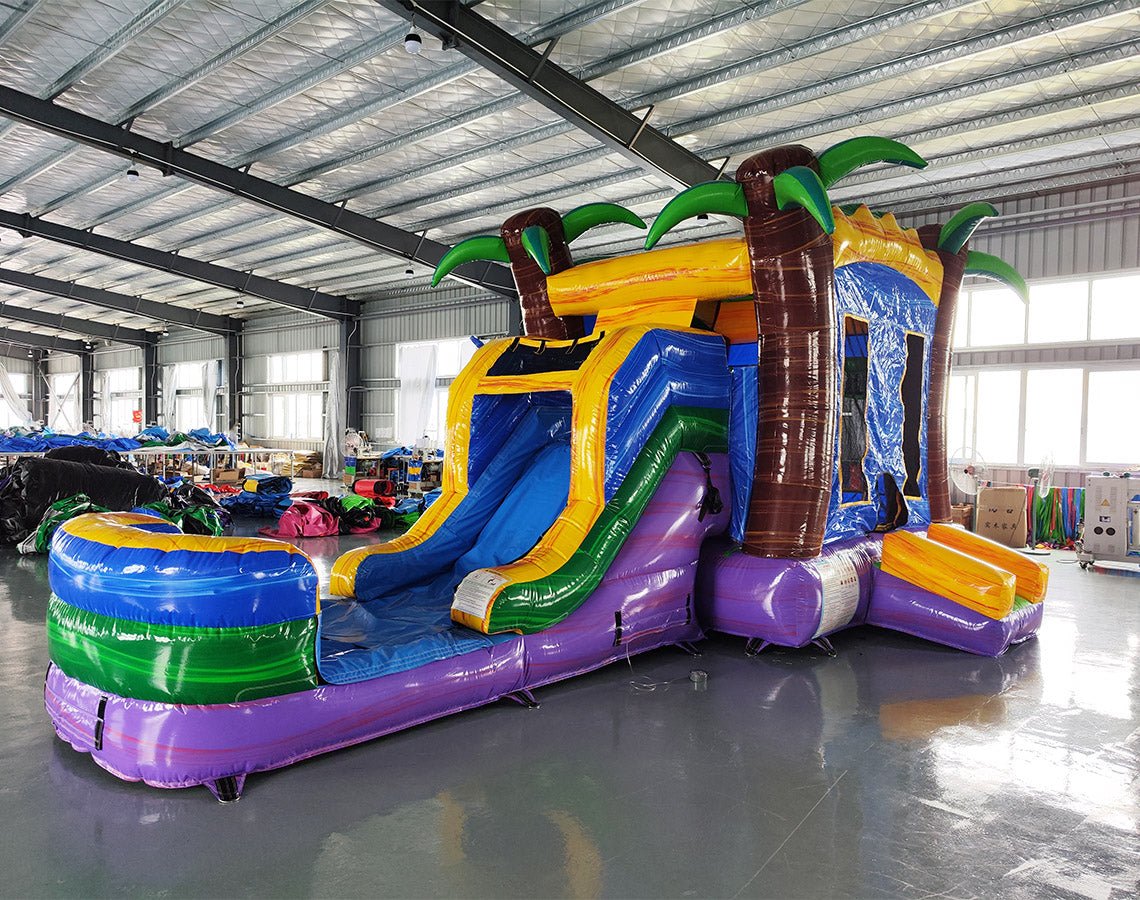 Wide-angle view of Goombay Splash Econo Combo showing bright blue, yellow, and purple design with inflatable palm trees, curved slide, and large splash pool area. The combo is set up in a spacious indoor warehouse with metal roofing, bright lighting, and other inflatable products visible in the background.