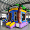 Side angle view of the Goombay Splash Econo Combo featuring a blue and yellow bounce house with palm tree decorations, curved slide, and spacious splash pool. The inflatable is set up in an indoor warehouse with other equipment and inflatables visible.