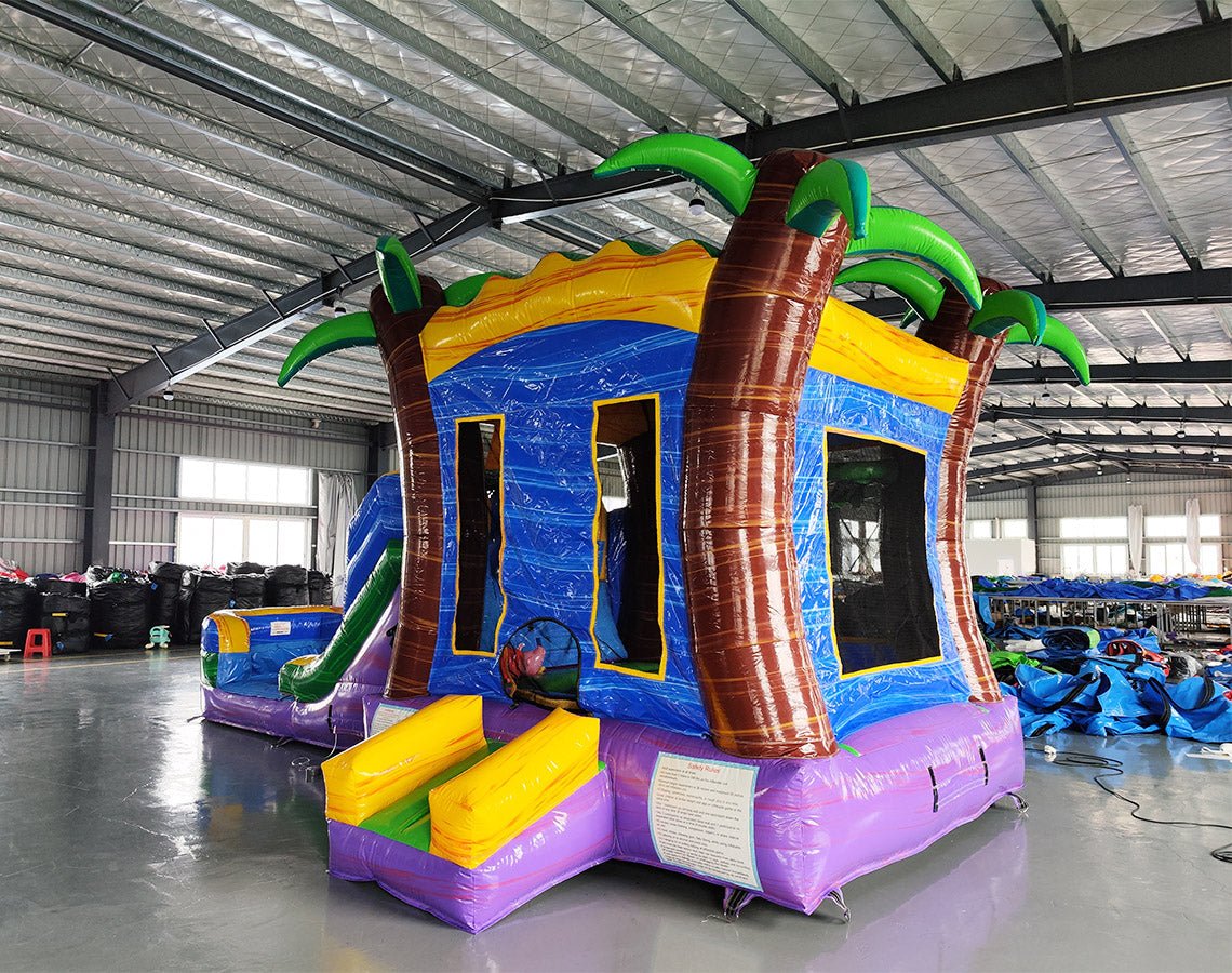 Side angle view of the Goombay Splash Econo Combo featuring a blue and yellow bounce house with palm tree decorations, curved slide, and spacious splash pool. The inflatable is set up in an indoor warehouse with other equipment and inflatables visible.