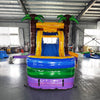 Large inflatable tropical-themed combo featuring a bounce house with palm tree decorations, attached slide, and splash pool. The structure has vibrant blue, yellow, and purple colors with brown palm tree trunks. Set up in an indoor warehouse with metal roofing and other inflatable products visible in the background.