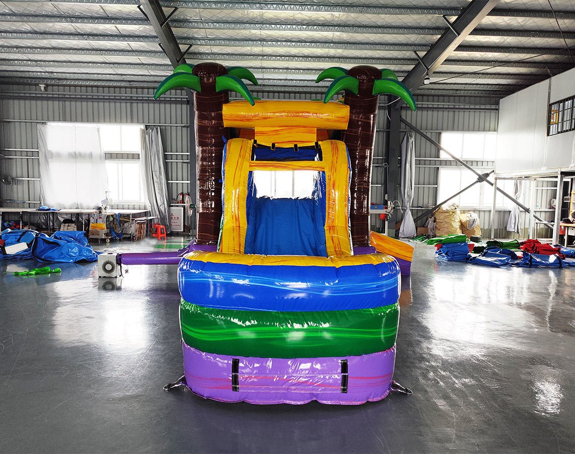 Large inflatable tropical-themed combo featuring a bounce house with palm tree decorations, attached slide, and splash pool. The structure has vibrant blue, yellow, and purple colors with brown palm tree trunks. Set up in an indoor warehouse with metal roofing and other inflatable products visible in the background.