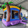 Front view of Goombay Splash Econo Combo, showcasing the bounce house entrance with palm tree decorations, blue slide, and colorful splash pool area. Set up in an indoor facility with other inflatable products and equipment visible in the background.