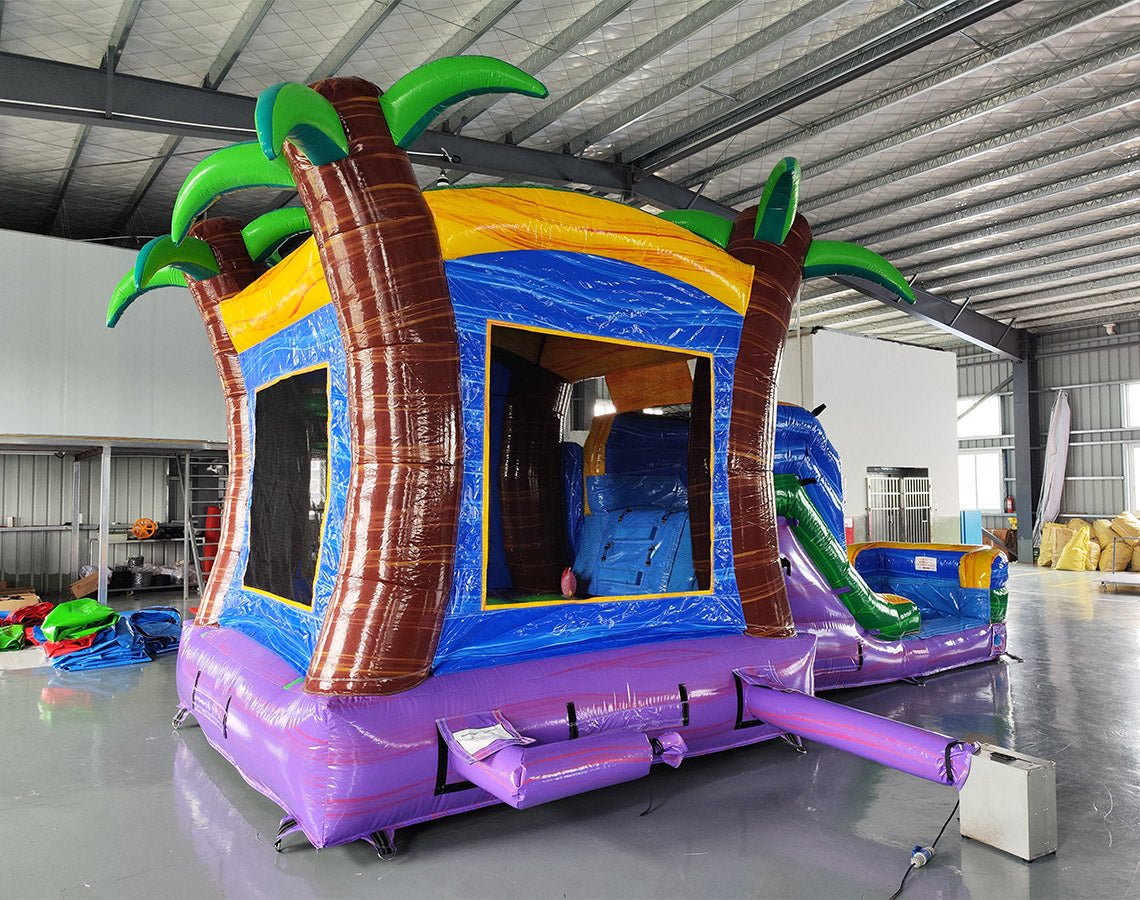 Front view of Goombay Splash Econo Combo, showcasing the bounce house entrance with palm tree decorations, blue slide, and colorful splash pool area. Set up in an indoor facility with other inflatable products and equipment visible in the background.