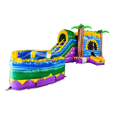 Large tropical-themed inflatable combo featuring dual curved blue slides flanked by inflatable palm trees. The structure includes a tall yellow climbing wall with blue accents, leading to the slide entrance. A spacious circular splash pool area with multicolored sections is visible at the base. Isolated on a white background, showcasing its vibrant design and wet/dry functionality.