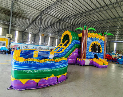 Wide-angle view of Goombay Curved Dual Lane Commercial Inflatable Combo (Wet/Dry) set up in a large indoor warehouse. Features dual blue curved slides, inflatable palm trees, and a tall yellow climbing structure. The large circular splash pool with multicolored sections is prominent in the foreground. Metal roofing, disco balls, and other warehouse equipment visible, providing context for its commercial use and size.