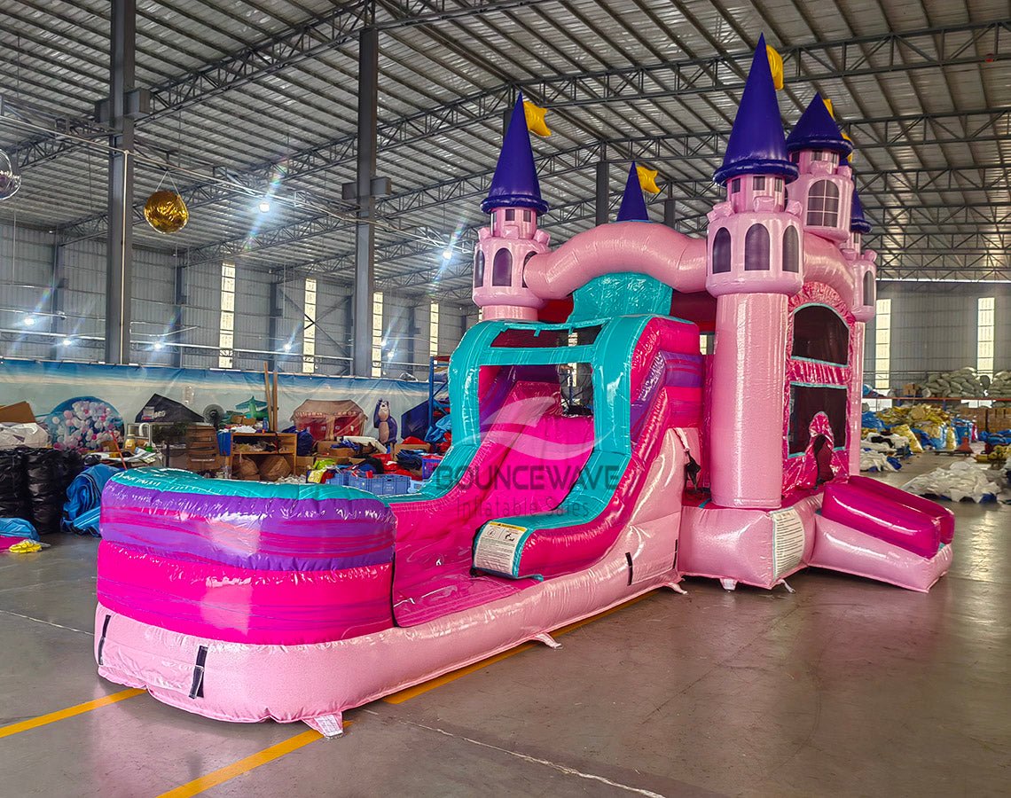 Side view of Grand Royal Castle Commercial Econo Inflatable Combo (Wet/Dry) set up in a large indoor warehouse. Features pink castle structure with purple turrets, yellow flags, teal slide, and pink splash pool. Metal roofing, disco ball, and other warehouse equipment visible, showcasing its size in a commercial setting.