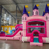 Front view of Grand Royal Castle Commercial Econo Inflatable Combo (Wet/Dry) in an indoor warehouse. Highlights the pink castle entrance with purple turrets, yellow flags, teal slide, and pink splash pool. Other inflatable products, storage containers, and warehouse equipment visible, demonstrating its scale in a commercial environment.