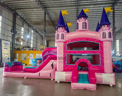 Front view of Grand Royal Castle Commercial Econo Inflatable Combo (Wet/Dry) in an indoor warehouse. Highlights the pink castle entrance with purple turrets, yellow flags, teal slide, and pink splash pool. Other inflatable products, storage containers, and warehouse equipment visible, demonstrating its scale in a commercial environment.