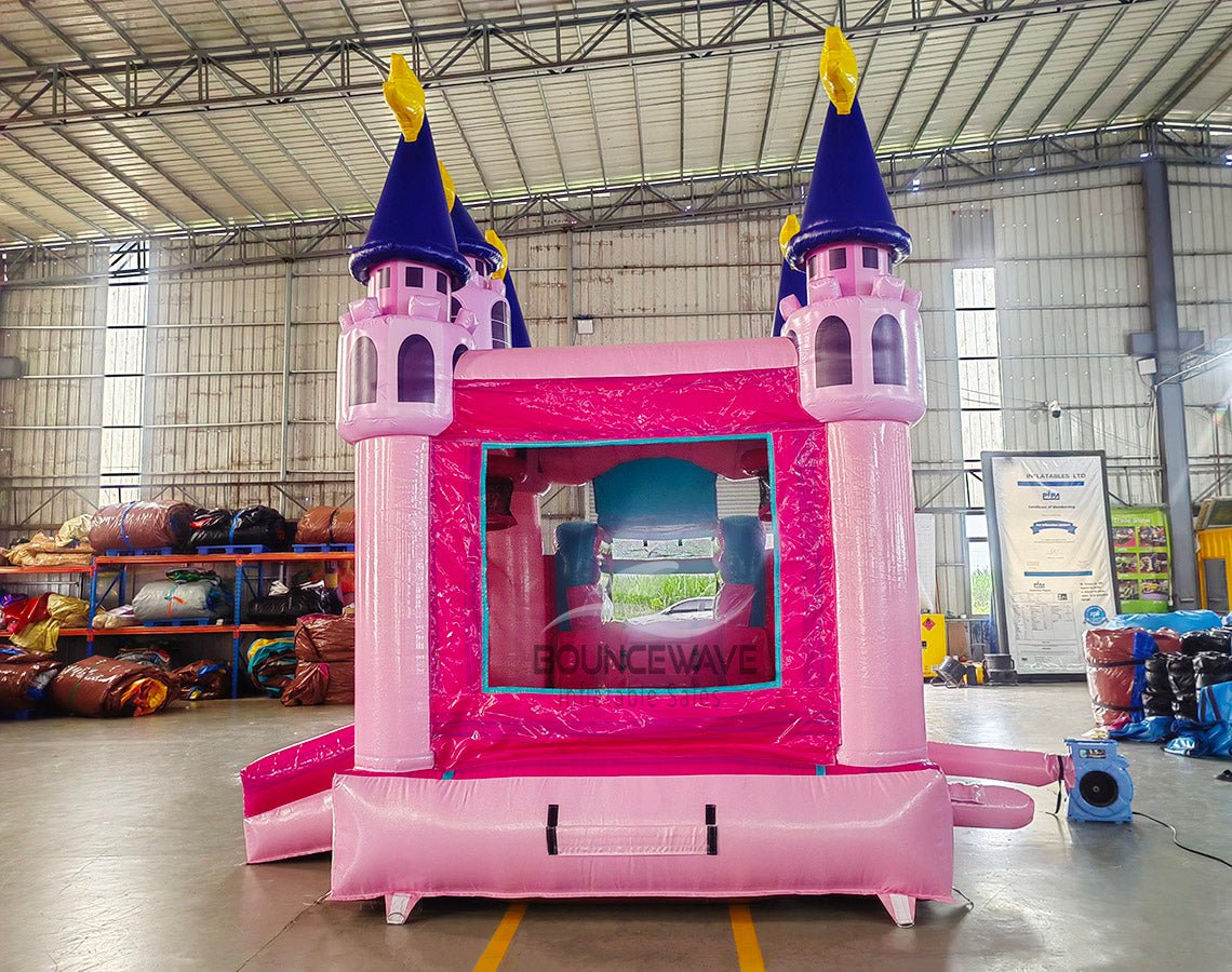 Close-up Rear view of Grand Royal Castle Commercial Econo Inflatable Combo (Wet/Dry) entrance. Shows detailed pink castle facade with purple turrets, yellow flags, and mesh-covered openings. Set up in a warehouse with other inflatable products and equipment visible in the background, emphasizing its design and commercial use.