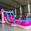 Wide-angle view of Grand Royal Castle Dual Lane Commercial Inflatable Combo in a spacious warehouse. The inflatable showcases a pink castle structure with purple turrets and yellow flags, dual slides in pink and blue, and a large splash pool area. Metal roofing, support beams, and storage shelves are visible, demonstrating its size and commercial setting.