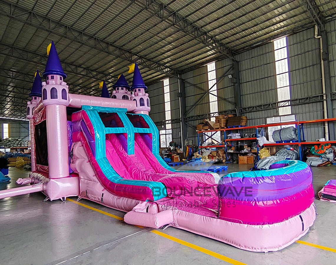 Wide-angle view of Grand Royal Castle Dual Lane Commercial Inflatable Combo in a spacious warehouse. The inflatable showcases a pink castle structure with purple turrets and yellow flags, dual slides in pink and blue, and a large splash pool area. Metal roofing, support beams, and storage shelves are visible, demonstrating its size and commercial setting.