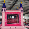 Rear view of Grand Royal Castle Dual Lane Commercial Inflatable Combo (Wet/Dry) set up in a large indoor warehouse. The inflatable features a pink castle structure with purple turrets topped with yellow flags. It includes dual slides in pink and blue, a bounce house area with mesh windows, and a splash pool. The "BOUNCEWAVE Inflatable Sales" logo is visible on the base. 