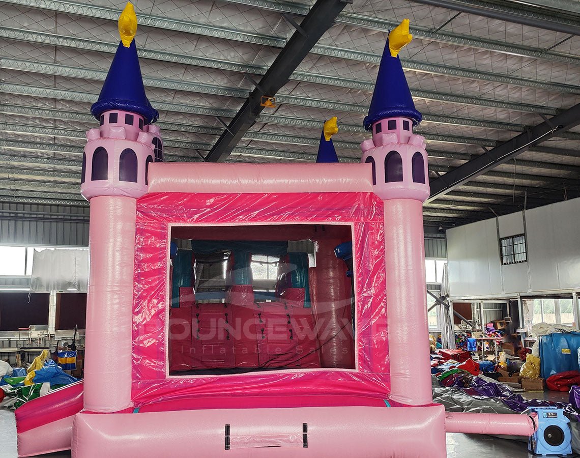 Rear view of Grand Royal Castle Dual Lane Commercial Inflatable Combo (Wet/Dry) set up in a large indoor warehouse. The inflatable features a pink castle structure with purple turrets topped with yellow flags. It includes dual slides in pink and blue, a bounce house area with mesh windows, and a splash pool. The "BOUNCEWAVE Inflatable Sales" logo is visible on the base. 