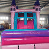 Front view of Grand Royal Castle Dual Lane Commercial Inflatable Combo set up in a large indoor warehouse. The inflatable features a pink castle structure with purple turrets, dual slides in pink and blue, and a splash pool area. The "BOUNCEWAVE Inflatable Sales" logo is visible on the base. Various other inflatable products and warehouse equipment are visible in the background.