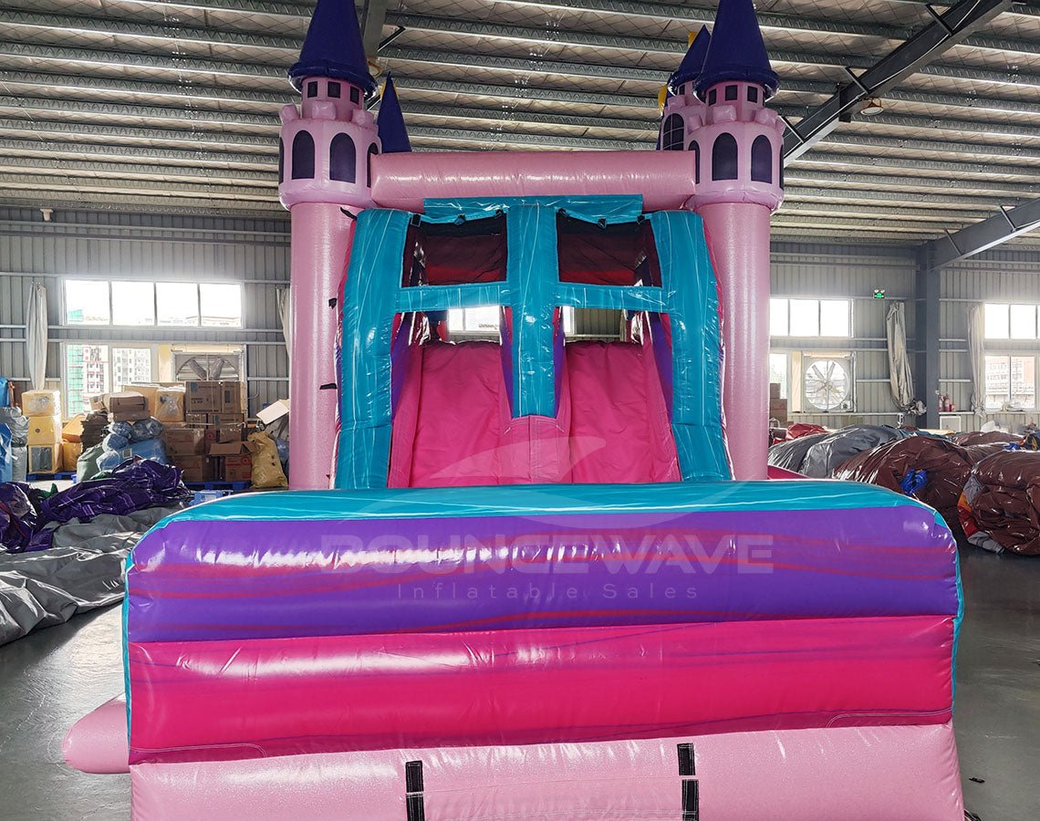 Front view of Grand Royal Castle Dual Lane Commercial Inflatable Combo set up in a large indoor warehouse. The inflatable features a pink castle structure with purple turrets, dual slides in pink and blue, and a splash pool area. The "BOUNCEWAVE Inflatable Sales" logo is visible on the base. Various other inflatable products and warehouse equipment are visible in the background.