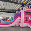 Three-quarter view of Grand Royal Castle Dual Lane Commercial Inflatable Combo in a warehouse environment. The inflatable features a pink castle structure with purple turrets and yellow flags, dual slides in pink and blue, a bounce house area, and a splash pool. Other inflatable products and warehouse materials in the background showcase its commercial application.