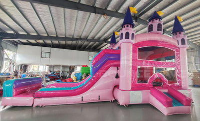 Three-quarter view of Grand Royal Castle Dual Lane Commercial Inflatable Combo in a warehouse environment. The inflatable features a pink castle structure with purple turrets and yellow flags, dual slides in pink and blue, a bounce house area, and a splash pool. Other inflatable products and warehouse materials in the background showcase its commercial application.