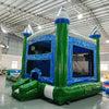 Three-quarter view of Green Gush Dual Lane Commercial Inflatable Combo in a warehouse setting. The inflatable displays green towers with blue and white striped tops, dual blue slides, a bounce house entrance, and a splash pool. Various other inflatable products and warehouse equipment are visible, demonstrating its size and commercial application.