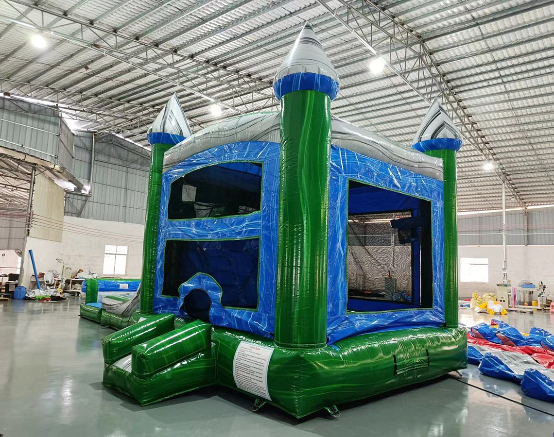 Three-quarter view of Green Gush Dual Lane Commercial Inflatable Combo in a warehouse setting. The inflatable displays green towers with blue and white striped tops, dual blue slides, a bounce house entrance, and a splash pool. Various other inflatable products and warehouse equipment are visible, demonstrating its size and commercial application.