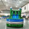 Front view of Green Gush Dual Lane Commercial Inflatable Combo featuring green towers with blue and white striped tops, dual blue slides, and a large splash pool. The structure includes a bounce house area with mesh windows. Set up in a spacious warehouse with various inflatable products and equipment visible in the background.