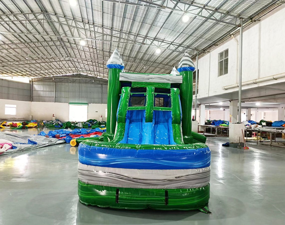 Front view of Green Gush Dual Lane Commercial Inflatable Combo featuring green towers with blue and white striped tops, dual blue slides, and a large splash pool. The structure includes a bounce house area with mesh windows. Set up in a spacious warehouse with various inflatable products and equipment visible in the background.