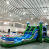 Wide-angle view of Green Gush Dual Lane Commercial Inflatable Combo set up in a large indoor facility. The structure features green towers with blue and white striped tops, dual blue slides, a bounce house area, and a spacious splash pool. The warehouse setting shows metal roofing, support beams, and other inflatable products, showcasing its commercial use context.