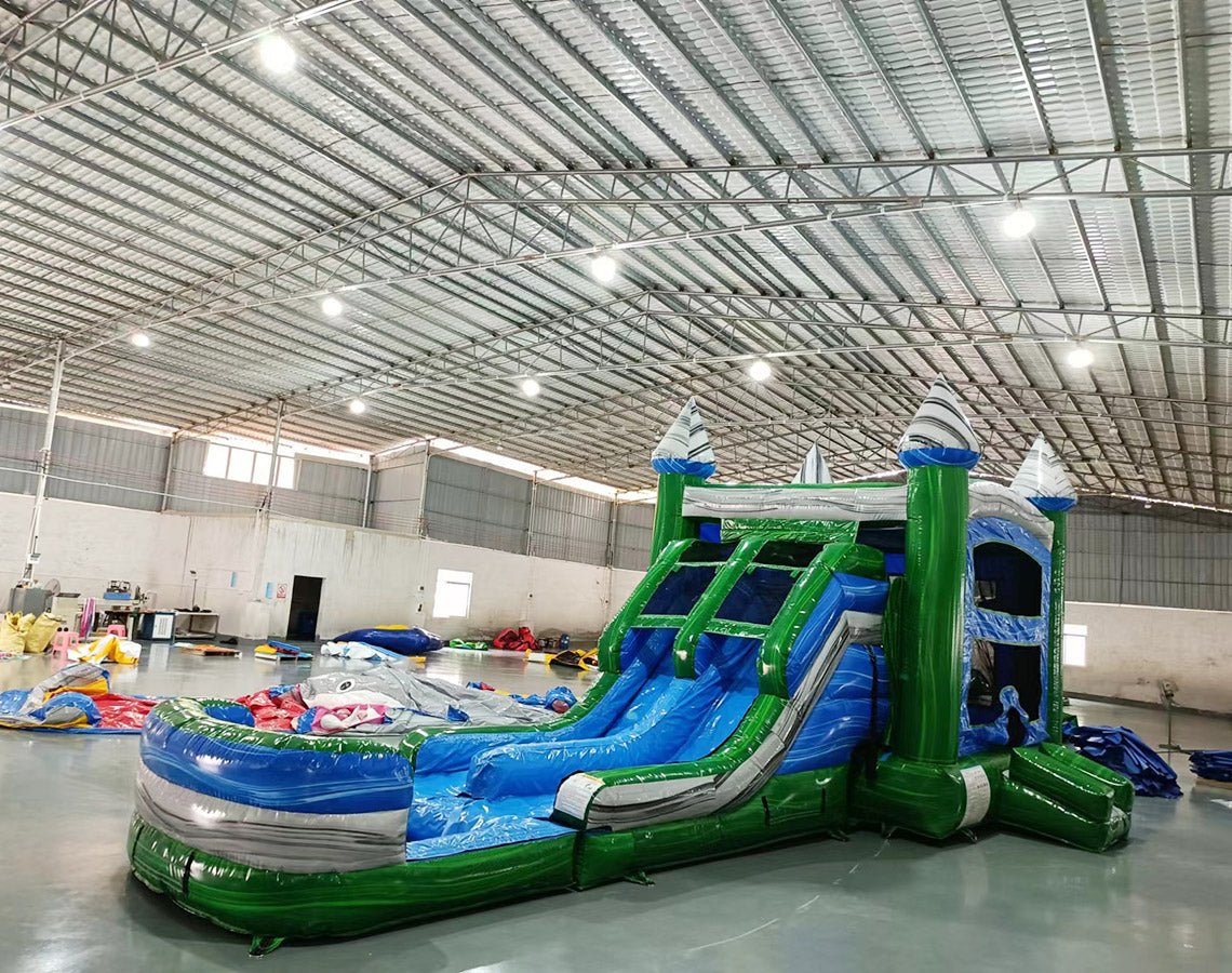 Wide-angle view of Green Gush Dual Lane Commercial Inflatable Combo set up in a large indoor facility. The structure features green towers with blue and white striped tops, dual blue slides, a bounce house area, and a spacious splash pool. The warehouse setting shows metal roofing, support beams, and other inflatable products, showcasing its commercial use context.