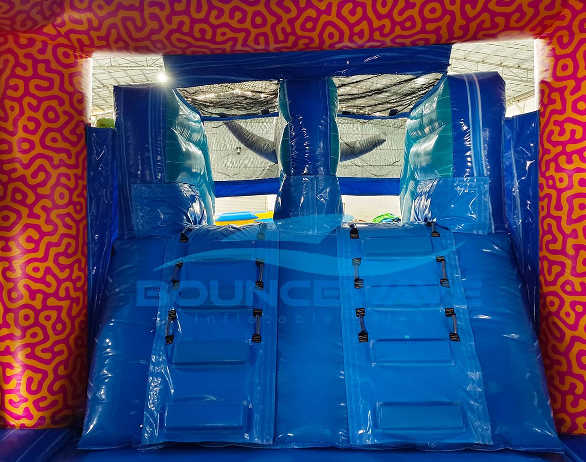 Interior view of Hammerhead Reef Dual Lane Commercial Inflatable Combo (Wet/Dry) showing dual blue slides with safety barriers. The inflatable features an underwater theme with blue walls and floor. Inflatable shark fins are visible at the top of the structure. The pink coral-patterned exterior frame is partially visible. Set against a backdrop of pink coral-patterned inflatable material, showcasing the combo's immersive design.