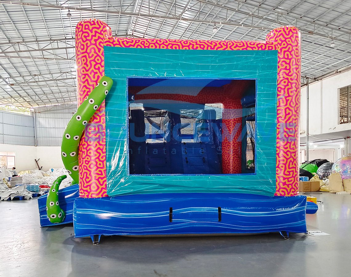 Rear view of Hammerhead Reef Dual Lane Commercial Inflatable Combo (Wet/Dry) featuring a vibrant ocean-themed design. The structure has a pink coral-patterned frame with a teal entrance, dual blue slides, and a large green octopus on top. Inflatable green tentacles with eyes flank the sides. A blue wave-patterned splash pool is visible at the base. Set up in a large indoor warehouse with metal roofing and various other inflatable products visible in the background.