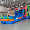 Front view of Hammerhead Reef Dual Lane Commercial Inflatable Combo featuring a vibrant ocean-themed design with dual blue slides, a large green octopus on top, and inflatable shark fins. The structure includes a pink coral-patterned bounce house area, mesh windows, and a splash pool. Set up in a spacious warehouse with metal roofing and other inflatable products visible in the background.