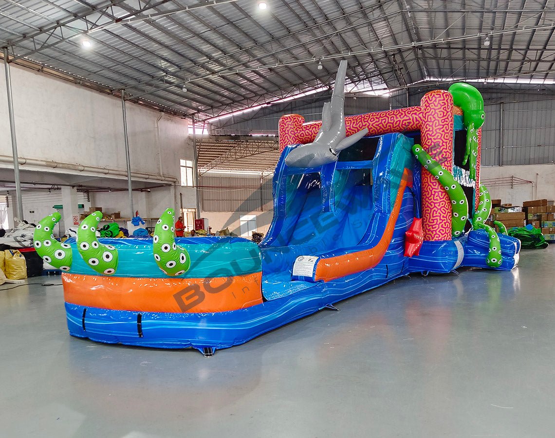 Front view of Hammerhead Reef Dual Lane Commercial Inflatable Combo featuring a vibrant ocean-themed design with dual blue slides, a large green octopus on top, and inflatable shark fins. The structure includes a pink coral-patterned bounce house area, mesh windows, and a splash pool. Set up in a spacious warehouse with metal roofing and other inflatable products visible in the background.