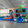 Wide-angle view of Hammerhead Reef Dual Lane Commercial Inflatable Combo in a large indoor facility. The inflatable displays dual blue slides, a green octopus on top, shark fins, and a pink coral-patterned bounce house. A splash pool and various sea-themed elements are visible. The warehouse setting shows metal roofing, support beams, and other inflatable products, demonstrating its size and commercial application.