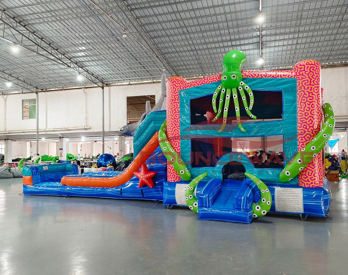 Wide-angle view of Hammerhead Reef Dual Lane Commercial Inflatable Combo in a large indoor facility. The inflatable displays dual blue slides, a green octopus on top, shark fins, and a pink coral-patterned bounce house. A splash pool and various sea-themed elements are visible. The warehouse setting shows metal roofing, support beams, and other inflatable products, demonstrating its size and commercial application.