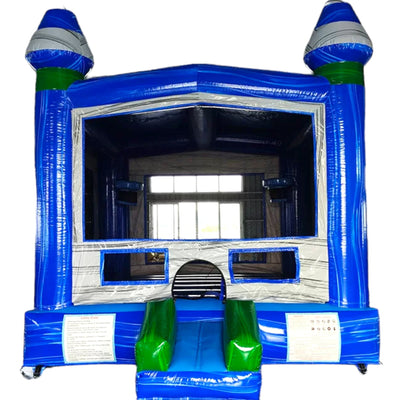 Large inflatable Island Drop Commercial Grade Bounce House featuring a vibrant blue and yellow design. The structure has a tall climbing wall with blue panels leading to two steep yellow slides. Inflatable palm trees flank the sides, adding a tropical theme. The bounce area has mesh windows for visibility and safety. Set up in a spacious indoor warehouse with metal roofing and fluorescent lighting. Other inflatable products and equipment are visible in the background, showcasing the commercial setting.