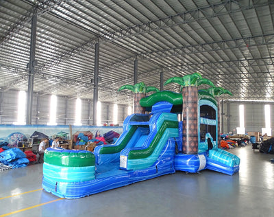 Wide-angle view of Island Drop Econo Combo showing bright blue and green design with inflatable palm trees, curved slide, and large splash pool area. The combo is set up in a spacious indoor warehouse with metal roofing, bright lighting, and other inflatable products visible in the background.