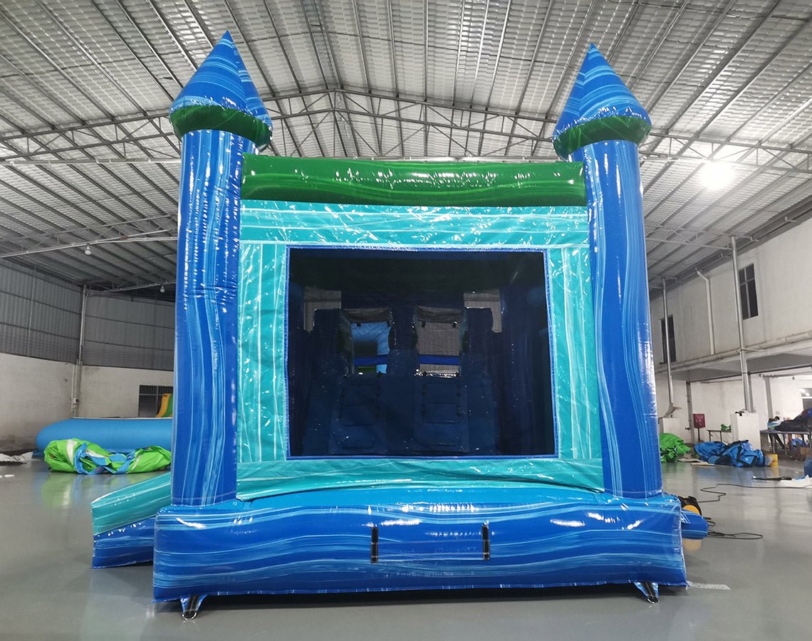 Rear view of Island Drop Splash & Save Dual Lane Inflatable Combo (Wet/Dry) set up in a large indoor warehouse. The inflatable features blue castle-like towers with conical tops, a turquoise bounce house area with mesh windows, and dual blue water slides. A green trim adorns the top, and a blue splash pool is visible at the base. The warehouse setting shows a metal roof with bright lighting, and various other inflatable products can be seen in the background.