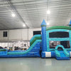 Wide-angle view of Island Drop Splash & Save Dual Lane Inflatable Combo set up in a large indoor warehouse. The inflatable features blue castle towers, dual water slides, and a turquoise bounce area. A blue splash pool is visible at the base. The warehouse setting shows metal roofing, bright lighting, and various other inflatable products in the background.