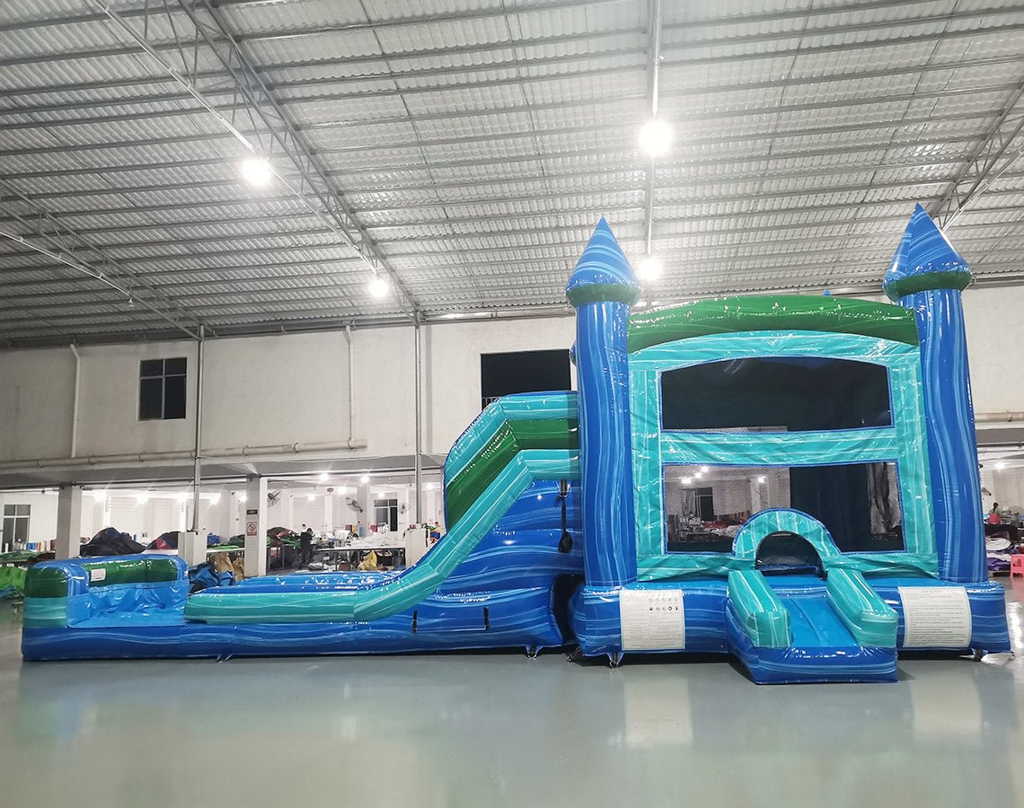 Wide-angle view of Island Drop Splash & Save Dual Lane Inflatable Combo set up in a large indoor warehouse. The inflatable features blue castle towers, dual water slides, and a turquoise bounce area. A blue splash pool is visible at the base. The warehouse setting shows metal roofing, bright lighting, and various other inflatable products in the background.