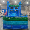 Front view of Island Drop Splash & Save Dual Lane Inflatable Combo showing the entrance area. Features blue castle towers, a turquoise bounce house frame with mesh windows, and a multi-colored splash pool base. The "BOUNCEWAVE Inflatable Sales" logo is prominently displayed. Set in a warehouse with various other inflatable products and equipment visible in the background.