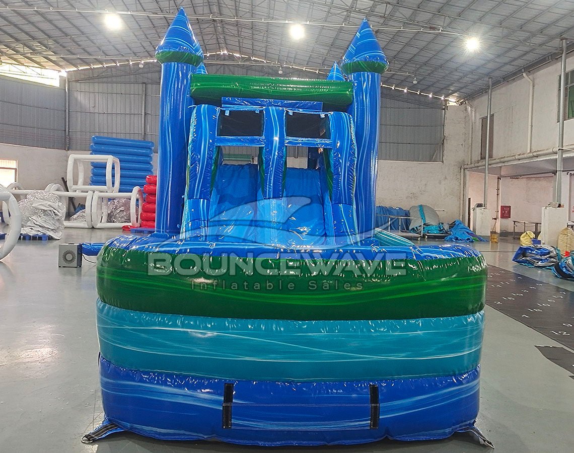 Front view of Island Drop Splash & Save Dual Lane Inflatable Combo showing the entrance area. Features blue castle towers, a turquoise bounce house frame with mesh windows, and a multi-colored splash pool base. The "BOUNCEWAVE Inflatable Sales" logo is prominently displayed. Set in a warehouse with various other inflatable products and equipment visible in the background.