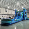 Three-quarter view of Island Drop Splash & Save Dual Lane Inflatable Combo in a spacious warehouse. The structure displays blue castle towers with conical tops, dual water slides, a turquoise bounce house, and a large blue splash pool. Other inflatable products and warehouse equipment are visible, demonstrating its size and commercial application.