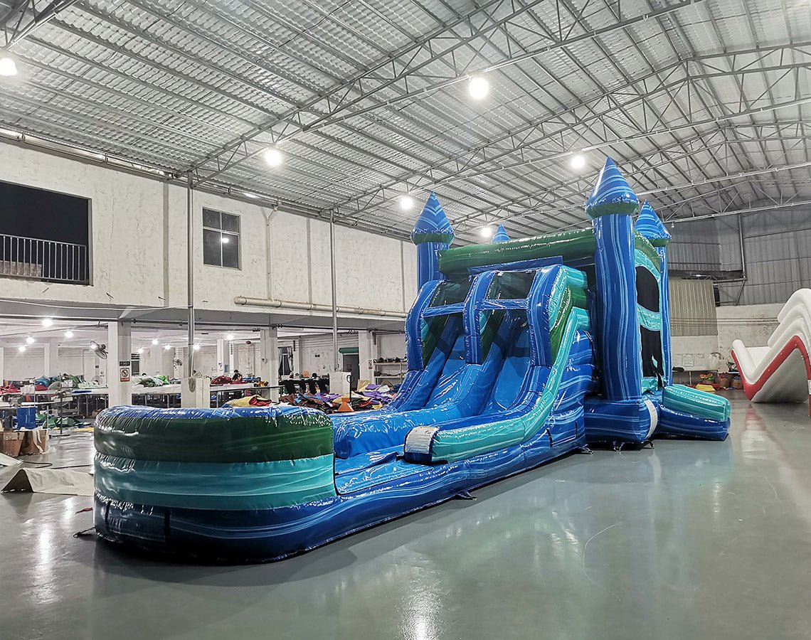 Three-quarter view of Island Drop Splash & Save Dual Lane Inflatable Combo in a spacious warehouse. The structure displays blue castle towers with conical tops, dual water slides, a turquoise bounce house, and a large blue splash pool. Other inflatable products and warehouse equipment are visible, demonstrating its size and commercial application.