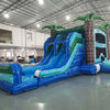 Wide-angle view of Island Drop Dual Lane Commercial Inflatable Combo in a large indoor facility. The inflatable displays dual blue water slides, green palm trees, a climbing wall, bounce area, and a spacious splash pool. The warehouse setting shows metal roofing, support beams, and other inflatable products, demonstrating its size and commercial application.