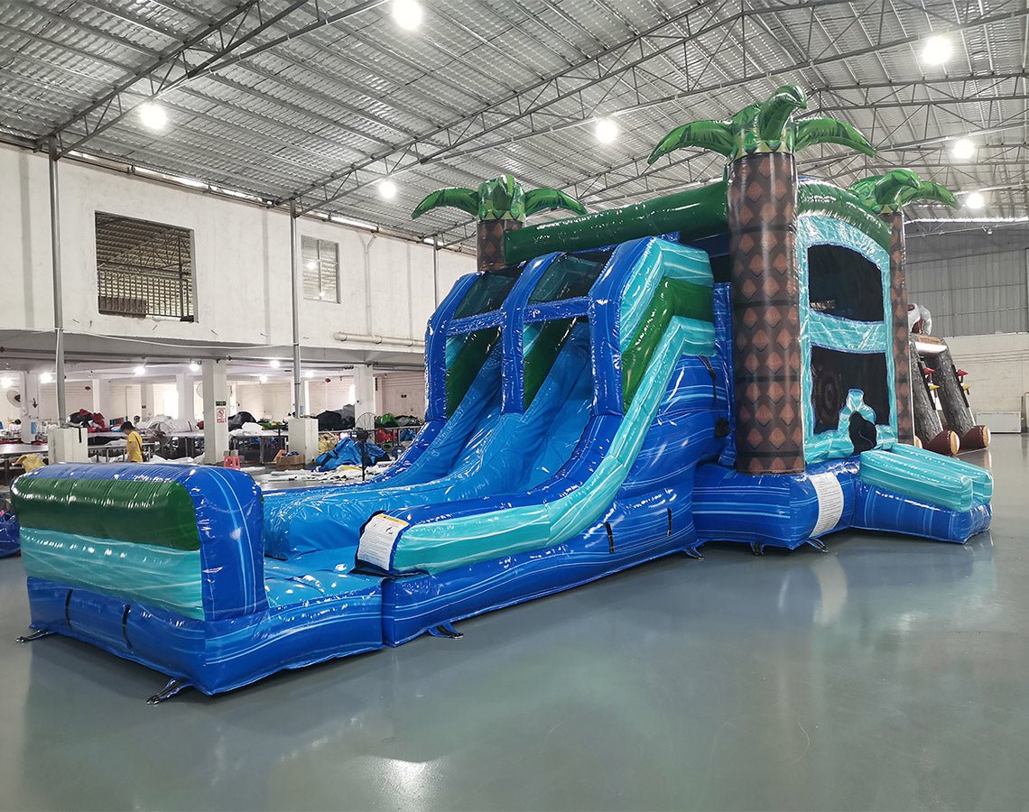 Wide-angle view of Island Drop Dual Lane Commercial Inflatable Combo in a large indoor facility. The inflatable displays dual blue water slides, green palm trees, a climbing wall, bounce area, and a spacious splash pool. The warehouse setting shows metal roofing, support beams, and other inflatable products, demonstrating its size and commercial application.