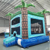 cross rear view of the Island Drop Dual Lane Commercial Inflatable Combo (Wet/Dry) focusing on the tropical-themed bounce house portion. Features a large inflatable palm tree pillar, mesh windows, and a blue base. Set up in a warehouse with concrete flooring, the image shows part of the climbing structure and slides in the background, emphasizing the combo's multi-functional design.