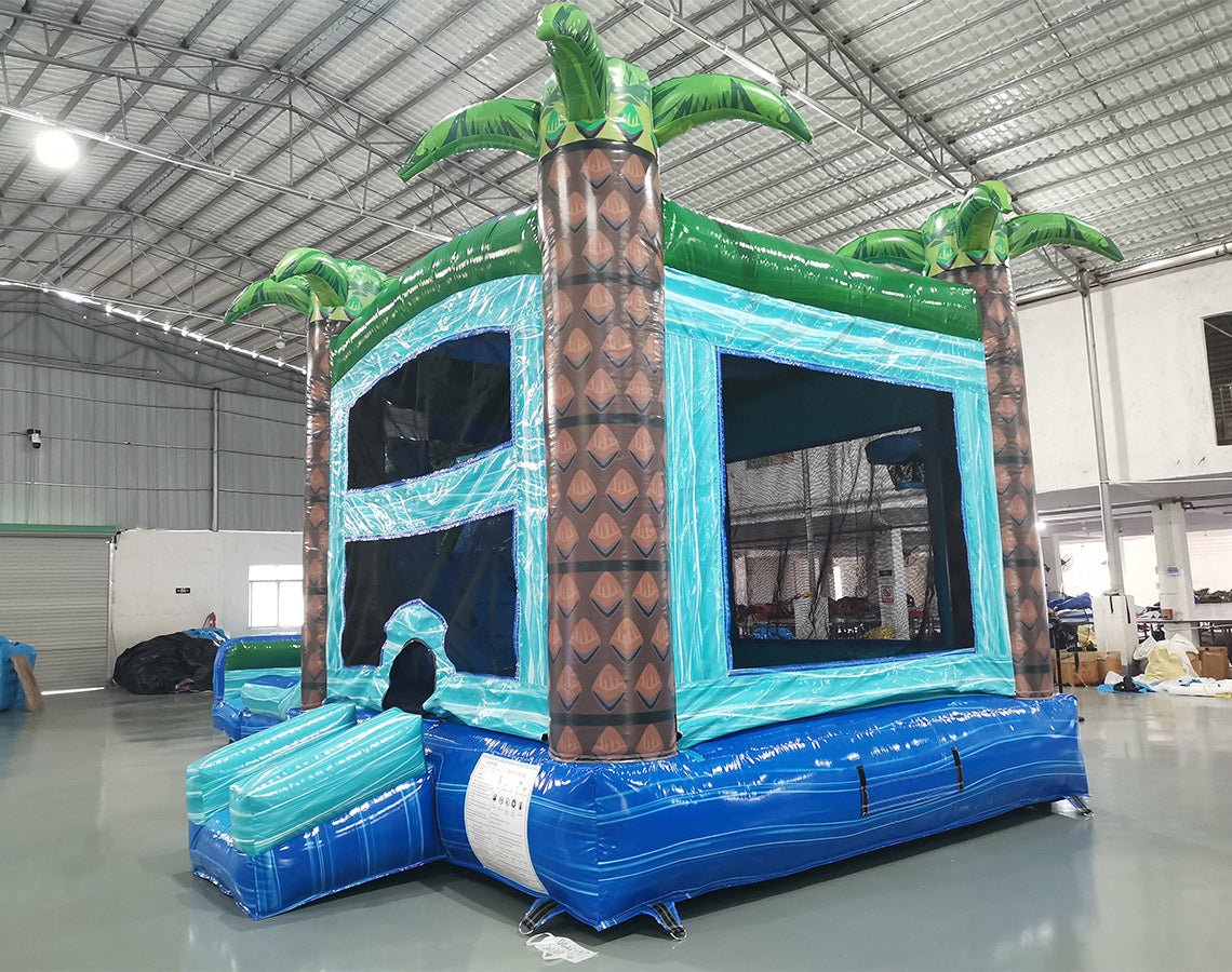 cross rear view of the Island Drop Dual Lane Commercial Inflatable Combo (Wet/Dry) focusing on the tropical-themed bounce house portion. Features a large inflatable palm tree pillar, mesh windows, and a blue base. Set up in a warehouse with concrete flooring, the image shows part of the climbing structure and slides in the background, emphasizing the combo's multi-functional design.