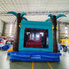 Close up Rear view of Island Drop Dual Lane Commercial Inflatable Combo (Wet/Dry) set up in a large indoor warehouse. Features a tropical-themed entrance with two large inflatable palm trees, a teal-colored frame, and a "BOUNCEWAVE" logo visible through the entrance. The base is blue with wave-like designs. Various equipment and materials are visible in the background, indicating a manufacturing or storage facility.