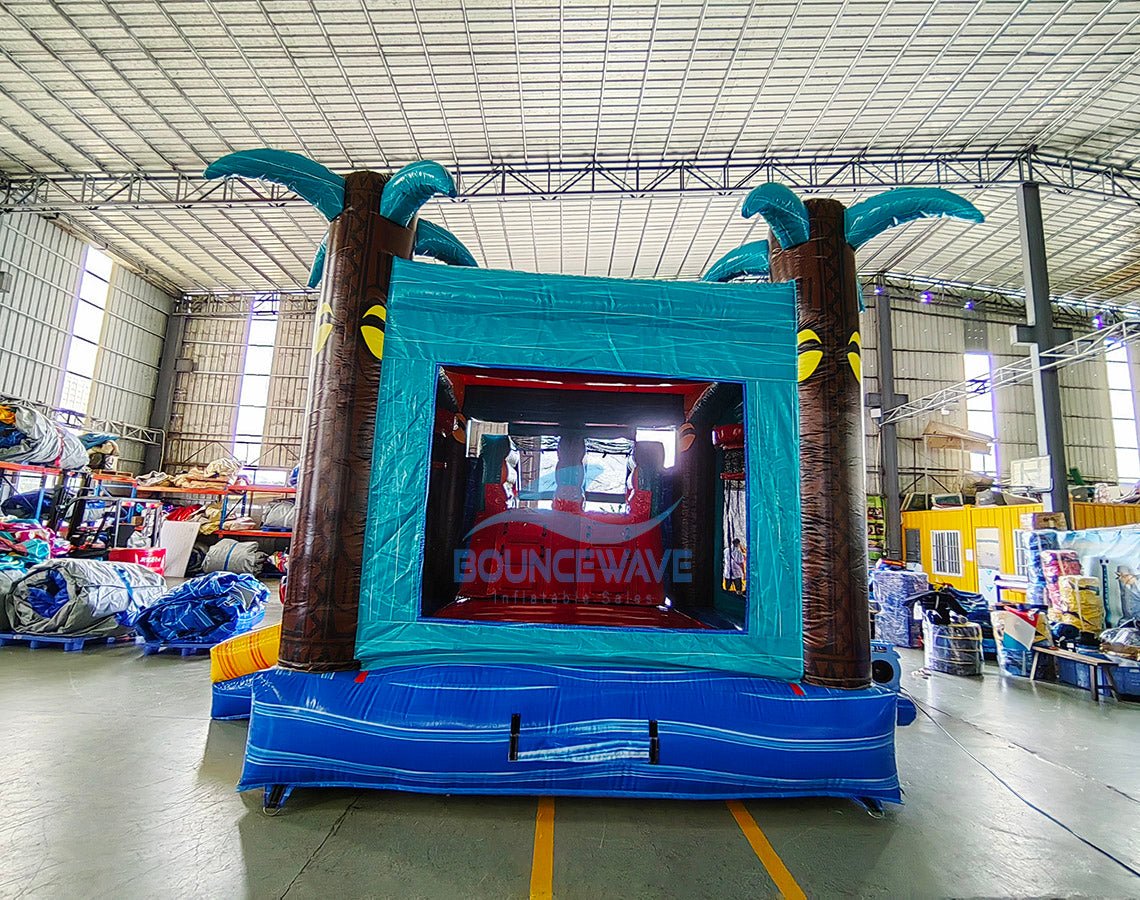 Close up Rear view of Island Drop Dual Lane Commercial Inflatable Combo (Wet/Dry) set up in a large indoor warehouse. Features a tropical-themed entrance with two large inflatable palm trees, a teal-colored frame, and a "BOUNCEWAVE" logo visible through the entrance. The base is blue with wave-like designs. Various equipment and materials are visible in the background, indicating a manufacturing or storage facility.