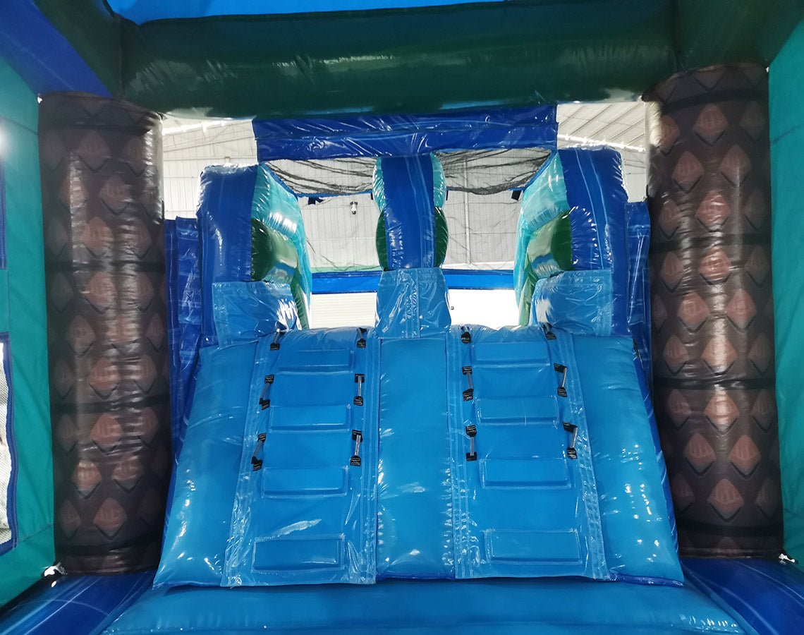 Interior view of Island Drop Dual Lane Commercial Inflatable Combo (Wet/Dry) showing two blue inflatable slides side by side. The slides are flanked by brown inflatable palm tree pillars with textured bark design. A blue inflatable base and teal-colored top frame are visible. The image captures the inside of the inflatable structure, showcasing its dual-lane design.